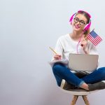 Uśmiechnieta młoda dziewczyna trzyma na kolanach laptopa, a w dłoni ma małą flagę USA