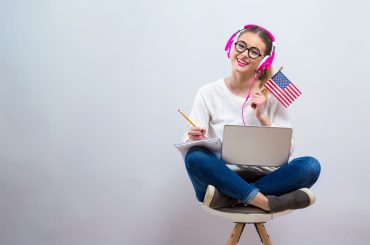 Uśmiechnieta młoda dziewczyna trzyma na kolanach laptopa, a w dłoni ma małą flagę USA